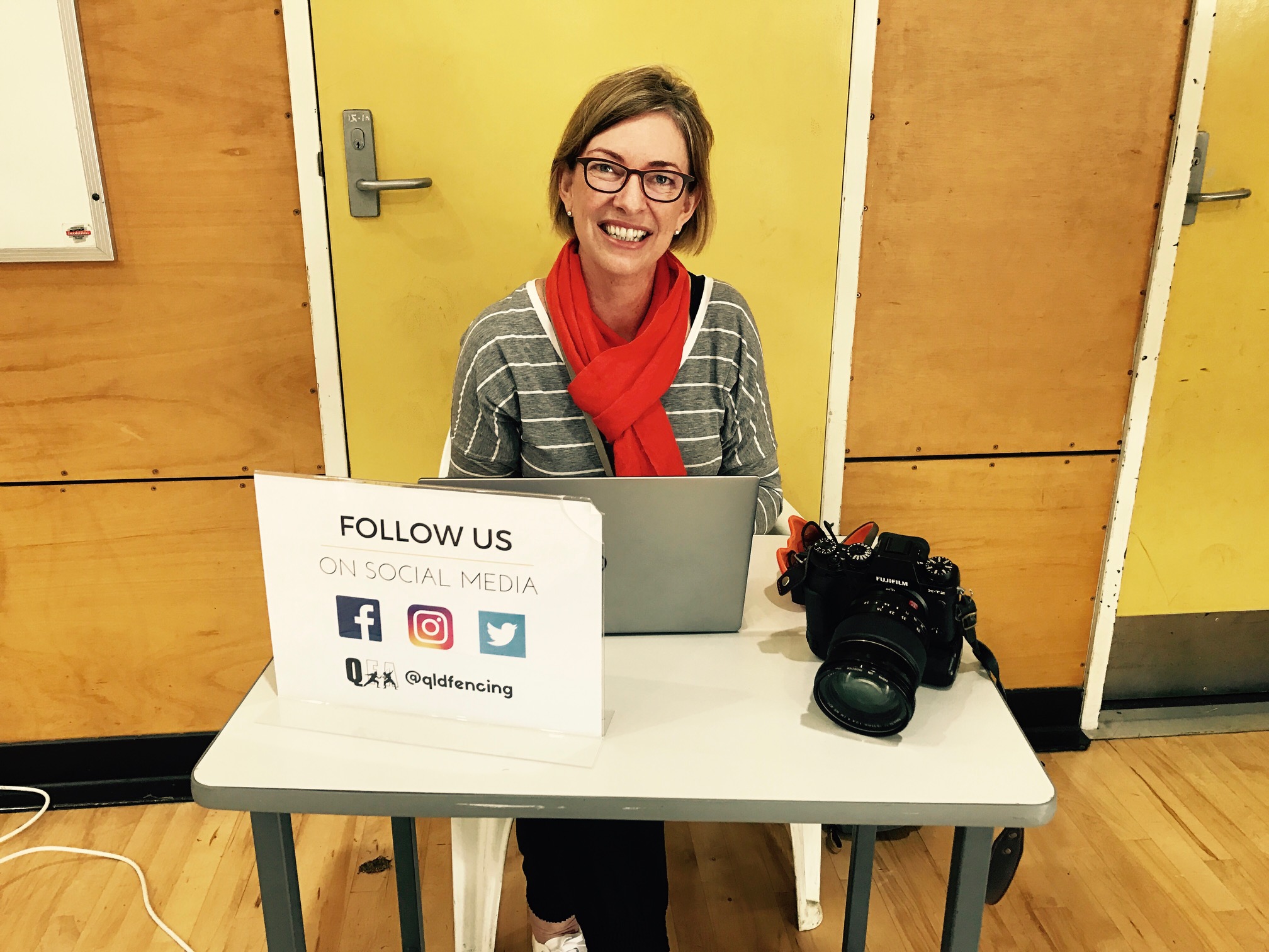 A woman wearing black-rimmed glasses and an orange scarf sits at a table behind an open laptop computer. There is a digital camera on the table and a sign with social media icons and the text 'follow us'. The woman is looking out and smiling.